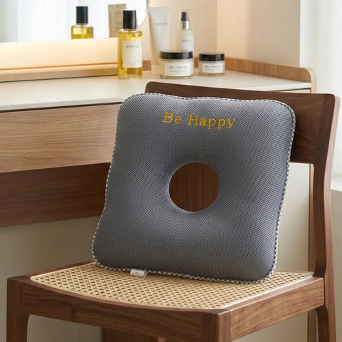 Gray donut pillow with 'Be Happy' text on a wooden chair, with skincare products on a vanity table in the background.