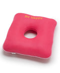 Pink square cushion with a circular hole and 'Be Happy' text on a white background