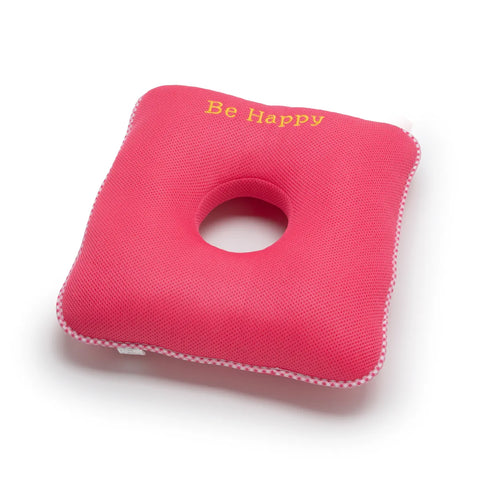 Pink square cushion with a circular hole and 'Be Happy' text on a white background