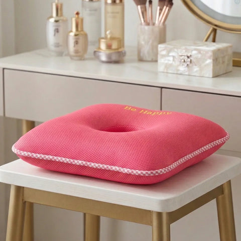 Pink square donut cushion with 'Be Happy' text on a white surface, surrounded by vanity items.