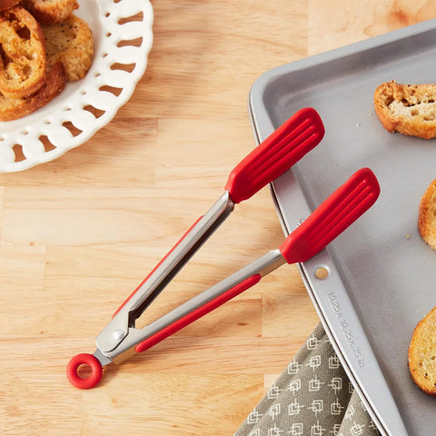 Red tongs on a wooden surface with a tray of bread slices.