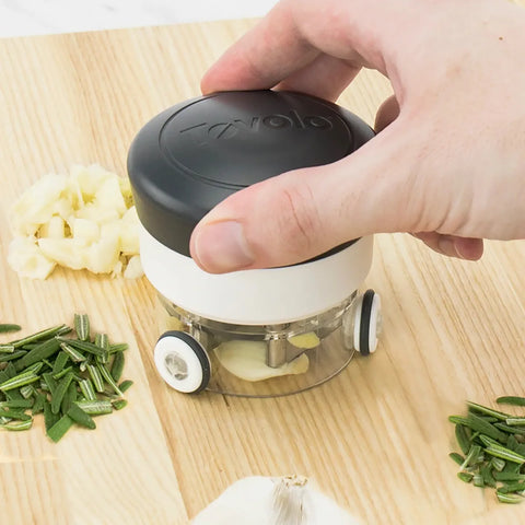 Hand using a garlic press on a wooden cutting board with chopped garlic and herbs.