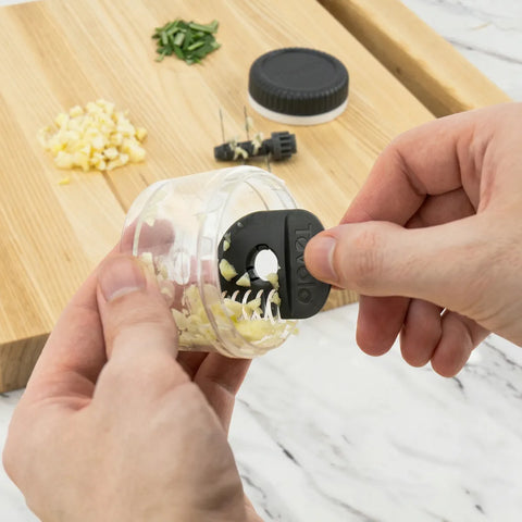 Person using a garlic chopper on a wooden cutting board with minced garlic and herbs.