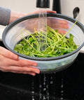 Person washing greens in a colander under running water in a kitchen.
