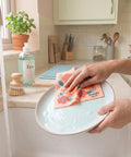 Person washing a dish with a pink dish cloth white orange and red poppy flowers over a kitchen sink with running water