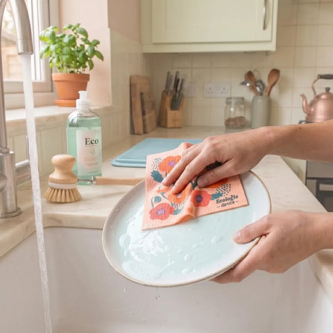 Person washing a dish with a pink dish cloth white orange and red poppy flowers over a kitchen sink with running water