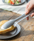 Person grabbing a slice of bread with metal tongs on a wooden table.
