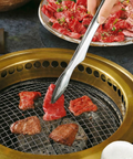 Grilling beef with metal tongs on a small grill with a plate of sliced beef in the background.