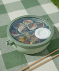 Round green bento box food container with different compartments with chopsticks on top of a green picnic blanket outdoors