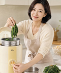 A woman in a beige cardigan and apron is using a cream-colored countertop food moisture remover in a bright kitchen, holding green vegetables above the steaming basket.