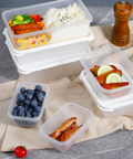Clear plastic food storage containers with white lids arranged on a beige cloth over a gray surface, shown with prepared meals, blueberries, sliced chicken, a pepper grinder, and a small tomato.