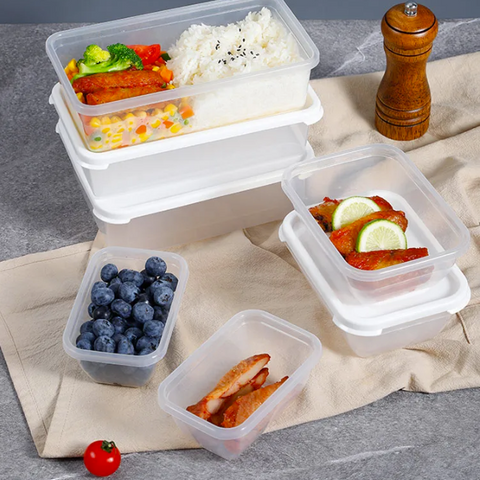 Clear plastic food storage containers with white lids arranged on a beige cloth over a gray surface, shown with prepared meals, blueberries, sliced chicken, a pepper grinder, and a small tomato.