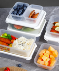 A set of clear plastic compartment food storage containers with white lids, shown filled with prepared meals and fruit on a gray stone surface. The containers include rice, broccoli, corn, tomatoes, blueberries, sliced watermelon, cantaloupe, and grilled fish with lime.