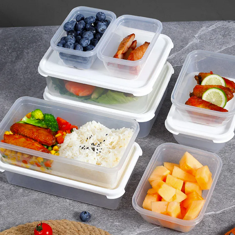 A set of clear plastic compartment food storage containers with white lids, shown filled with prepared meals and fruit on a gray stone surface. The containers include rice, broccoli, corn, tomatoes, blueberries, sliced watermelon, cantaloupe, and grilled fish with lime.