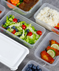 Clear plastic meal prep containers filled with various foods, including a salad with lettuce, tomatoes, blueberries, shrimp, and a piece of meat; white rice with black sesame seeds; fried chicken with lime slices; and other meals, arranged on a gray stone-textured surface.