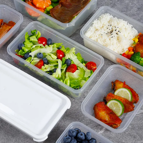 Clear plastic meal prep containers filled with various foods, including a salad with lettuce, tomatoes, blueberries, shrimp, and a piece of meat; white rice with black sesame seeds; fried chicken with lime slices; and other meals, arranged on a gray stone-textured surface.