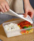 A clear plastic bento lunch box filled with white rice, glazed chicken, broccoli, and mixed vegetables, placed on a woven round mat with a person lifting the white lid.