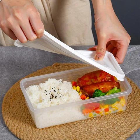 A clear plastic bento lunch box filled with white rice, glazed chicken, broccoli, and mixed vegetables, placed on a woven round mat with a person lifting the white lid.