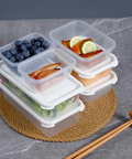 A stack of clear plastic meal prep containers with white lids, shown on a woven round mat with food samples including blueberries, salad, salmon, and fruit slices. Chopsticks rest nearby on a gray stone surface.