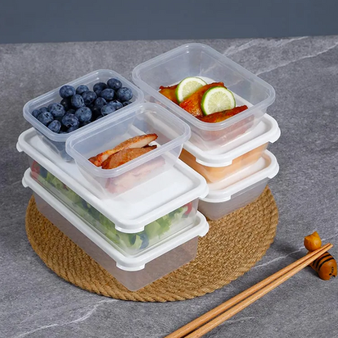 A stack of clear plastic meal prep containers with white lids, shown on a woven round mat with food samples including blueberries, salad, salmon, and fruit slices. Chopsticks rest nearby on a gray stone surface.