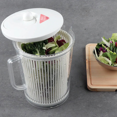 Salad spinner with fresh greens on a gray surface, next to a bowl of salad.