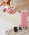 Person holding a pink bottle with a cap, next to a pink bottle on a drying rack.