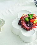 A white ceramic plate with a flower-shaped rim holds a plated octopus salad with tomatoes, green beans, and parsley garnish, set on a white tablecloth next to a matching small dish of mixed beans and peas.