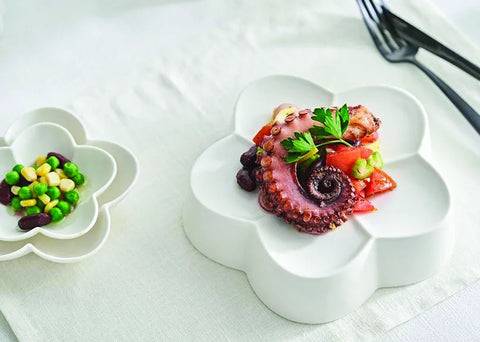 A white ceramic plate with a flower-shaped rim holds a plated octopus salad with tomatoes, green beans, and parsley garnish, set on a white tablecloth next to a matching small dish of mixed beans and peas.
