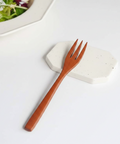 A wooden pasta fork with a warm reddish-brown finish rests diagonally on a white speckled ceramic coaster, with a blurred plate of salad in the background.