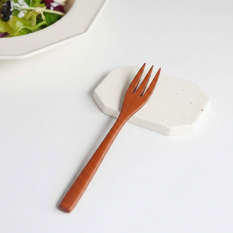 A wooden pasta fork with a warm reddish-brown finish rests diagonally on a white speckled ceramic coaster, with a blurred plate of salad in the background.