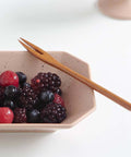 A light wood two-pronged fork rests across a speckled beige ceramic bowl filled with mixed berries, set on a clean white surface.