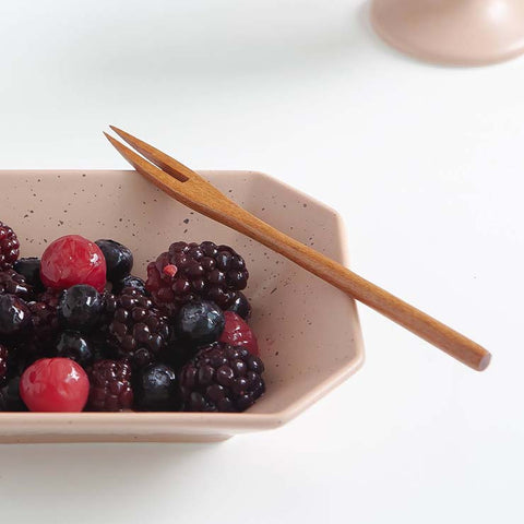 A light wood two-pronged fork rests across a speckled beige ceramic bowl filled with mixed berries, set on a clean white surface.
