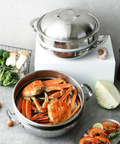 A stainless steel multi-tier steaming pot set displayed on a gray stone surface with food props. The top pot with lid sits on a white block, while the lower pot contains cooked crab meat and carrots.