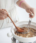 A close-up of a person's hands holding a wooden spoon and a metal strainer over a white ceramic pot on a stove, cooking a brown mushroom soup or stew.