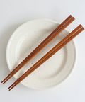 A pair of brown wooden chopsticks resting diagonally on a round off-white ceramic plate with subtle dark speckles, shot from a top-down perspective on a clean white background.