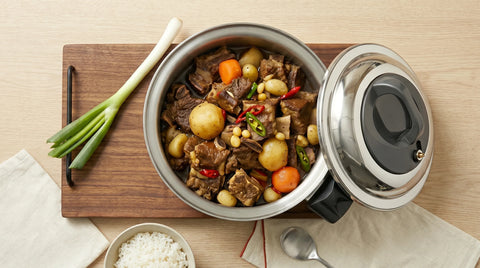 Stew in a pot on a wooden cutting board with vegetables and a spoon on a light wood background