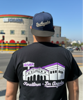 A man viewed from behind wearing a black t-shirt and a navy blue baseball cap. The back of the t-shirt features a graphic of the Koreatown & Los Angeles building with purple and white details.