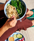 Person eating a bowl of noodles with greens, wearing green sandals on a sidewalk.