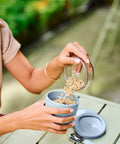 Person pouring granola from a container into a bowl on a table outdoors.