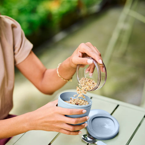 Person pouring granola from a container into a bowl on a table outdoors.