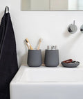 Two matte dark gray cylindrical toothbrush holders sit on a white bathroom sink next to a black towel hanging on a wall hook. One holder contains toothbrushes, and a matching gray soap dish with a red hair tie sits nearby.