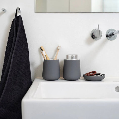 Two matte dark gray cylindrical toothbrush holders sit on a white bathroom sink next to a black towel hanging on a wall hook. One holder contains toothbrushes, and a matching gray soap dish with a red hair tie sits nearby.